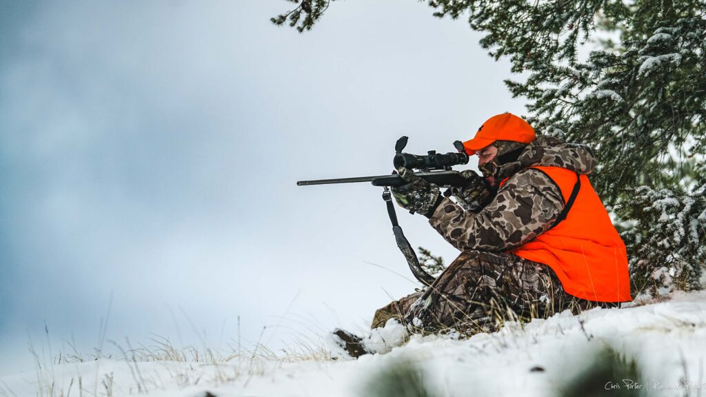 Hunter wearing orange aiming a rifle while sitting on a hill in the snow with trees at his back and blue sky to the left.